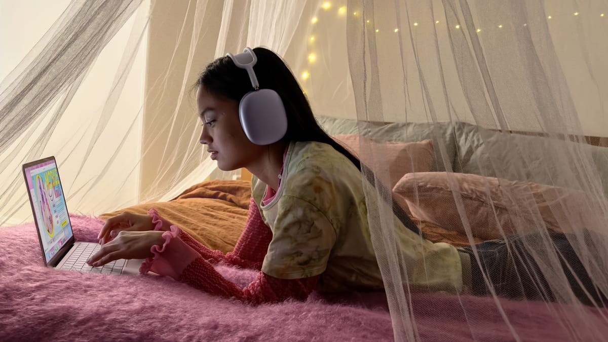 A young child with MacBook Neo in pink and AirPods Max (which cost almost as much as the laptop) in purple on a pretty fuzzy blanket in their bed