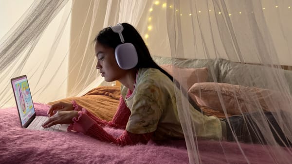 A young child with MacBook Neo in pink and AirPods Max (which cost almost as much as the laptop) in purple on a pretty fuzzy blanket in their bed